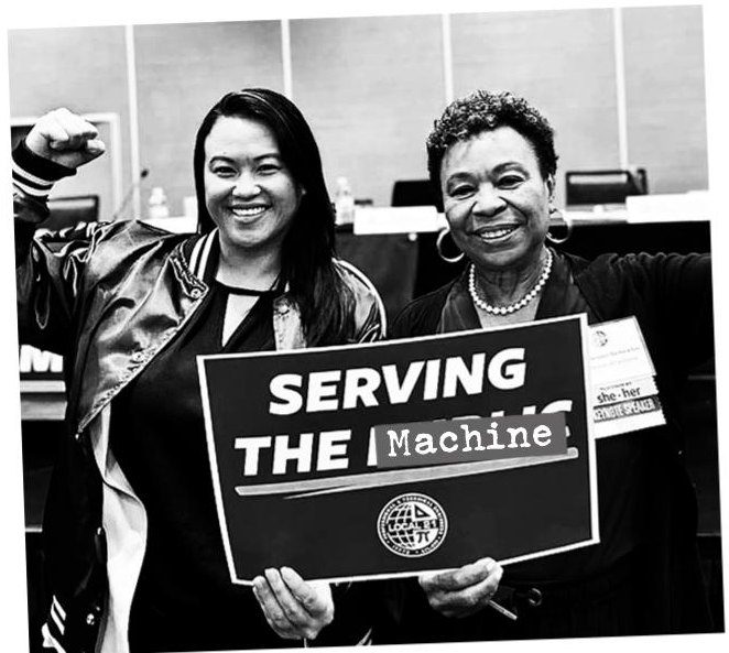 Barbara Lee, fist raised, beside a supporter holding a sign altered to read "Serving the Machine".
